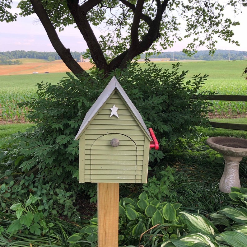 Op de afbeelding: Een groene houten brievenbus met een witte ster aan de voorkant en een rode hendel. De brievenbus is gemonteerd op een houten paal en is omgeven door groene planten.