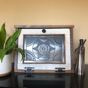 May include: A white and brown wooden bread box with a tin panel door featuring a starburst design. The door has two black metal hinges and a black knob.