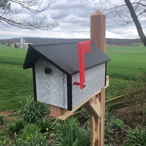 May include: A gray and black mailbox with a red flag attached to a wooden post. The mailbox has a black roof and a white, textured front. The post is made of natural wood.