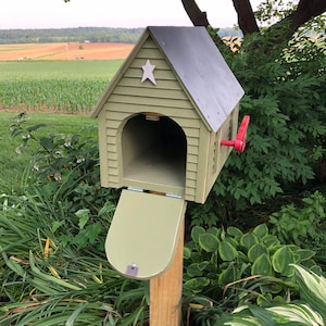 Op de afbeelding: Een groene houten brievenbus met een ster aan de voorkant en een rode vlag. De brievenbus is open en de deur hangt naar beneden. De brievenbus staat op een houten paal en is omgeven door groene planten.