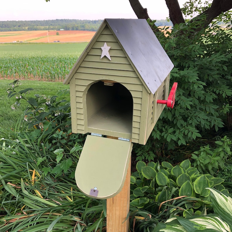 Op de afbeelding: Een groene houten brievenbus met een ster aan de voorkant en een rode vlag. De brievenbus is open en de deur hangt naar beneden. De brievenbus staat op een houten paal en is omgeven door groene planten.