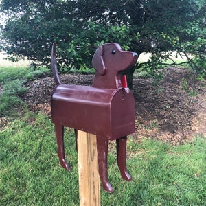 Puede incluir: Un buz&oacute;n de color marr&oacute;n con forma de perro y una solapa roja. El buz&oacute;n est&aacute; montado en un poste de madera.