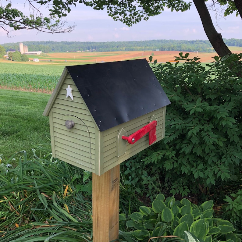 Op de afbeelding: Een groene houten brievenbus met een zwart dak en een rode vlag. De brievenbus is gemonteerd op een houten paal en is omgeven door groen gras en planten.