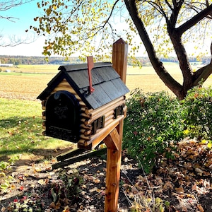 May include: A wooden log cabin style mailbox with a black metal door and a red flag. The mailbox is mounted on a wooden post and is surrounded by fallen leaves.