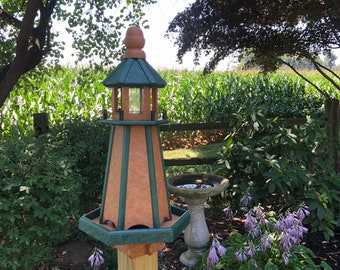 May include: A lighthouse-shaped bird feeder with a brown and green color scheme. The feeder is mounted on a wooden post and has a small window. The bird feeder is surrounded by greenery, a bird bath, and a wooden fence.