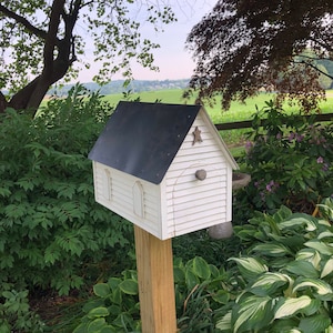 Op de afbeelding: Een wit vogelhuisje met een zwart dak en een ster aan de voorkant. Het vogelhuisje is gemonteerd op een houten paal en is omgeven door groen gebladerte.