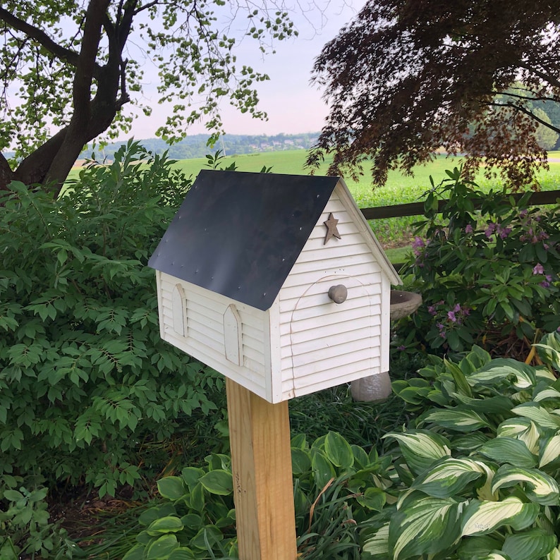 Op de afbeelding: Een wit vogelhuisje met een zwart dak en een ster aan de voorkant. Het vogelhuisje is gemonteerd op een houten paal en is omgeven door groen gebladerte.