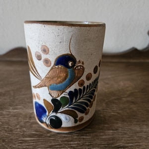 May include: A ceramic tumbler with a bird design. The off-white tumbler features a hand-painted bird with blue, brown, and teal accents. The bird is surrounded by brown dots and blue and green foliage. The rim and base are brown.
