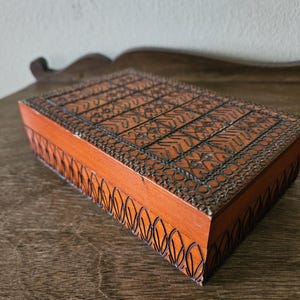 May include: A rectangular wooden box with intricate carved patterns on the lid and sides. The box is stained a warm reddish-brown color, with dark accents in the carved details. The design features geometric and floral motifs.