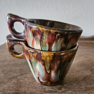 May include: Two stacked ceramic mugs with a unique, organic shape. The mugs feature a rich, earthy color palette of brown, red, yellow, and green, with a glossy finish. Each mug has a handle and a wide, shallow bowl.