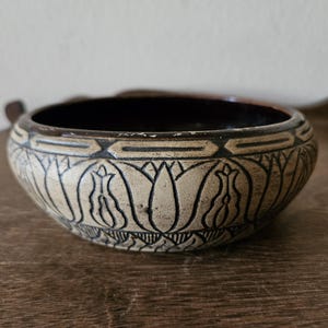 May include: A round ceramic bowl with a cream-colored exterior featuring a dark brown incised floral pattern and a geometric border around the rim. The interior of the bowl is finished in a dark brown glaze, and the overall design has a textured appearance.