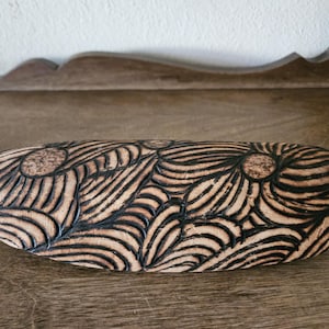 May include: A wooden eyeglasses case with a floral design. The case is oval-shaped and features a carved pattern of stylized flowers and leaves. The wood has a light brown color with dark brown accents in the carved details.