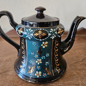 May include: A black teapot with a teal and gold painted design. The teapot has a round body with a flared spout and a handle.