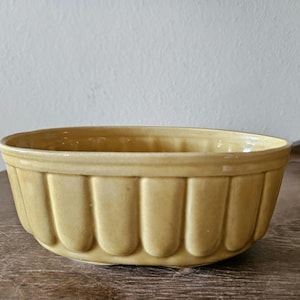 May include: A light yellow, oval-shaped ceramic bowl. The bowl has a scalloped design on the sides and a smooth rim. The bowl is sitting on a wooden surface.