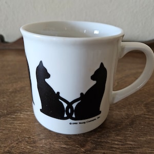 May include: White ceramic mug with black silhouette of two cats facing each other. The mug has a handle and the text "© 1990 Young Concepts, Inc." on the bottom.