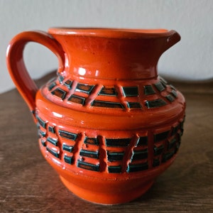 May include: Orange ceramic pitcher with a geometric pattern in black and green. The pitcher has a wide mouth and a handle.