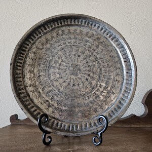 May include: A silver tray with intricate engraved designs, resting on a black metal stand. The tray has a circular shape and a raised rim.