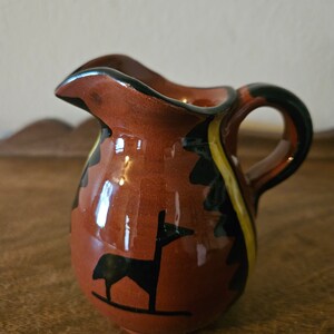 May include: A small, brown ceramic pitcher with a black and yellow geometric design. The pitcher has a handle and a spout. A black silhouette of a stylized animal is painted on the front.