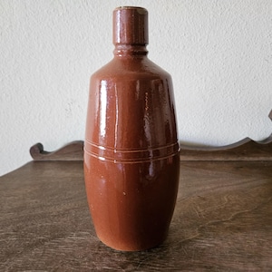 May include: A brown ceramic bottle with a narrow neck and a rounded body. The bottle has a glossy finish and two horizontal bands around the middle. It is displayed on a wooden surface.