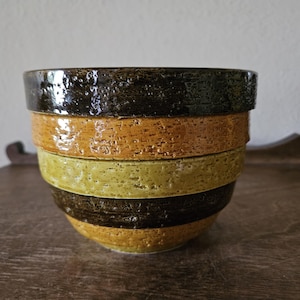 May include: A round ceramic bowl featuring a horizontal striped pattern with alternating bands of dark brown, mustard yellow, and olive green. The exterior surface has a distinct, rough, and textured finish throughout the entire bowl.
