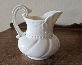 May include: A white ceramic creamer with a decorative handle and spout. The creamer has a bulbous body with a spiral design and a beaded band around the middle. The handle has a scrolled design. The creamer is approximately 5 inches tall.