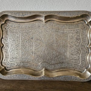 May include: Rectangular silver-toned tray with ornate floral and geometric patterns. The tray has a scalloped edge and a raised rim. The design includes a Star of David in the center. A decorative serving piece.