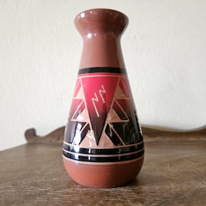Peut inclure: Un vase en terre cuite avec une base évasée et un col étroit. Le vase présente un motif géométrique en rouge, noir et beige. Le motif comprend des triangles et des zigzags. Le vase est posé sur une surface en bois.