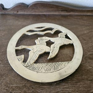 May include: Circular brass trivet featuring a detailed relief of two flying ducks over a grassy area. The trivet has a decorative border and is designed to protect surfaces from hot dishes. A vintage kitchen accessory.