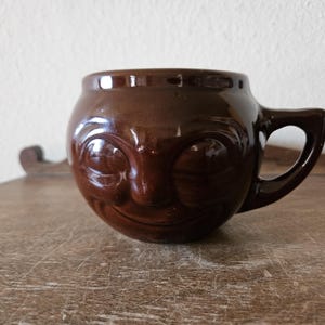 May include: A brown ceramic mug with a smiling face design. The mug has a rounded body, a handle, and a glossy finish. The mug is sitting on a wooden surface.