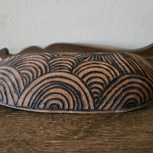 May include: A wooden bowl with a carved design of black waves. The bowl is oval shaped and has a natural brown color.