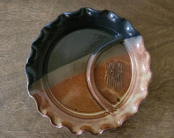 May include: A handcrafted ceramic serving dish with a unique design. The dish features a divided section with a wavy rim. The colors include black, brown, and tan. The dish has a textured, carved design in the smaller section.