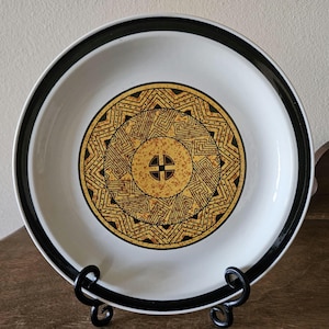 May include: A white ceramic dinner plate with a black rim and a gold and black geometric design in the center.