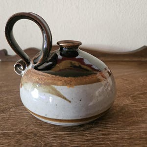 May include: A handcrafted ceramic pitcher with a rounded body and a looped handle. The pitcher features a white base with brown and gold accents, and a dark brown handle and rim. The design is simple and elegant, suitable for serving beverages.