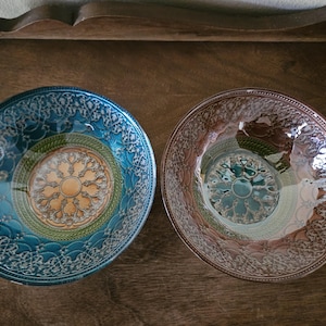 May include: Two decorative glass bowls with detailed patterns. One is teal with an orange and gold central design, the other light brown. Both bowls have an ornate border. The bowls are likely for serving.
