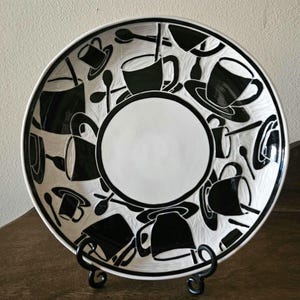 May include: A white ceramic plate with a black coffee-themed design. The plate features black illustrations of coffee cups, spoons, and saucers around the rim, with a large white circle in the center. The plate is displayed on a black metal stand.