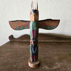 May include: A hand-carved wooden totem pole featuring an owl design. The totem pole has a natural wood base and is painted with various colors, including brown, green, red, and black. The wings are painted with a detailed pattern. The totem pole is approximately 8 inches tall.