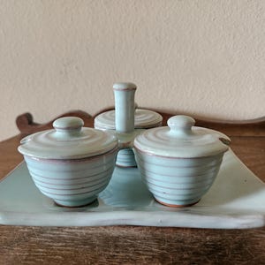 May include: A light blue ceramic condiment set with two lidded bowls and a central handle, resting on a matching rectangular tray. The bowls and lids have horizontal lines and a subtle pink rim. The set is designed for serving condiments.