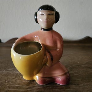May include: A ceramic planter shaped like a woman in a pink robe holding a yellow pot.