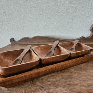 May include: A wooden serving set featuring three square bowls and small spoons, all crafted from a warm-toned wood. The bowls are set on a rectangular tray, perfect for serving snacks or dips.