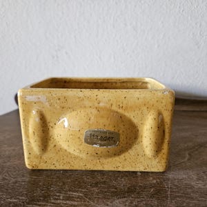 May include: A rectangular, speckled yellow ceramic container with rounded edges. The container has an oval indentation on the front and a small label that reads "Haeger". The container is sitting on a wooden surface.
