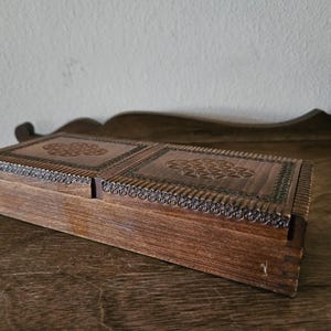 May include: A rectangular wooden box with a hinged lid, featuring intricate floral carvings on the top and sides. The box is stained a warm brown color and appears to be vintage or antique. The box is resting on a wooden surface.
