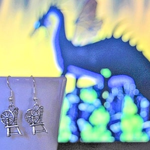 May include: Silver-colored earrings featuring a spinning wheel design. The earrings are detailed with a chair-like base and a wheel. The background is a blurred image with yellow, blue, and green hues.