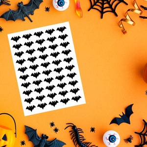 May include: A sheet of white paper covered in black bat stickers, surrounded by Halloween decorations on an orange background. Decorations include plastic spiders, bats, a jack-o'-lantern, and a spider web.