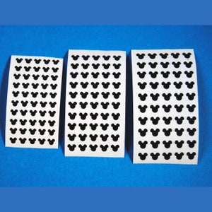 May include: Three white sticker sheets with black Mickey Mouse head shapes arranged in rows. The sheets are of varying sizes, with the largest sheet containing the most stickers. The stickers are on a blue background.