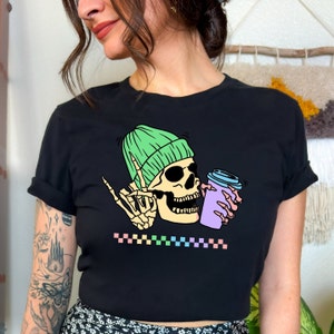 May include: Black t-shirt with a graphic of a skeleton wearing a green beanie and holding a purple coffee cup. The graphic has a rainbow checkered pattern underneath.