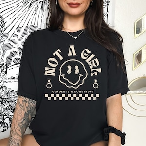 May include: Black t-shirt with a white graphic of a smiley face and the text "NOT A GIRL" in a circle. The text "GENDER IS A CONSTRUCT" is below the circle, and a checkered pattern is below the text.