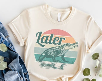 May include: A cream colored t-shirt with a vintage style graphic of an alligator in a swamp with the word "Later" above it.
