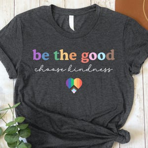 Może przedstawiać: Szary t-shirt z napisem "be the good choose kindness" w kolorach tęczy. Pod napisem znajduje się tęczowe serce z białym symbolem pokoju.