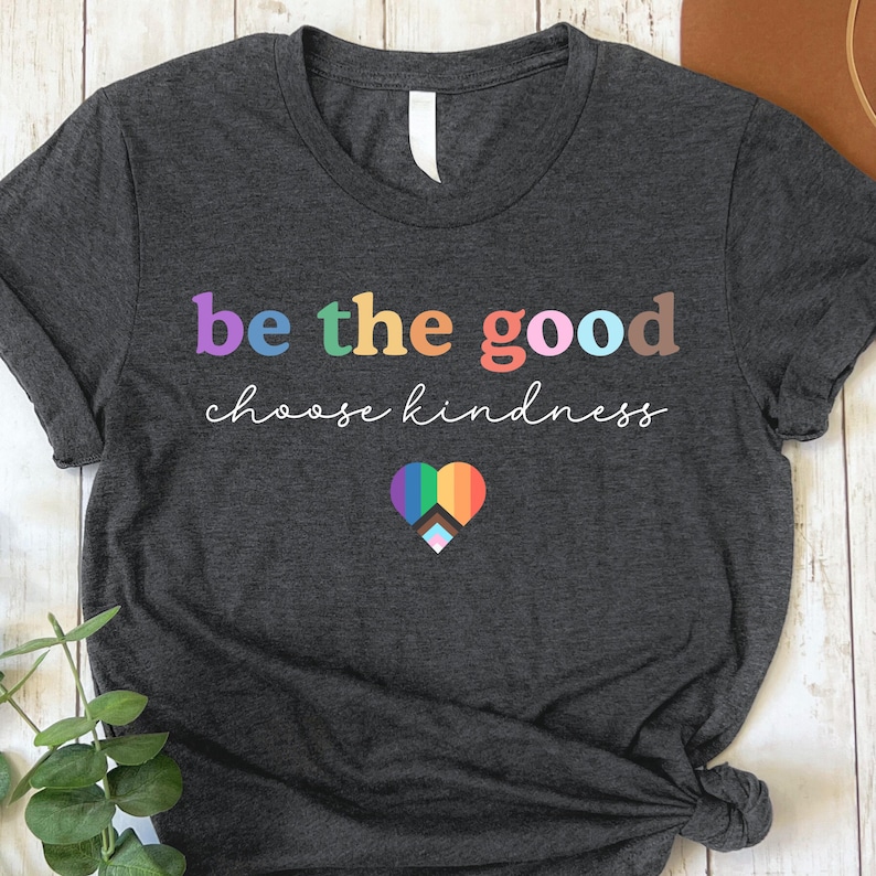 Może przedstawiać: Szary t-shirt z napisem "be the good choose kindness" w kolorach tęczy. Pod napisem znajduje się tęczowe serce z białym symbolem pokoju.