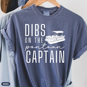 May include: A blue t-shirt with white text that reads "Dibs on the pontoon captain" and a white outline of a pontoon boat.